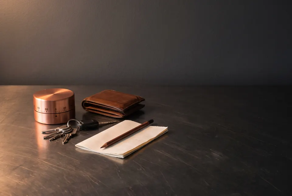 Scène générée : une minuterie de cuisine en cuivre, un portefeuille de cuir fermé, des clés d'auto et une note vierge avec un crayon sur une table d'acier foncé.