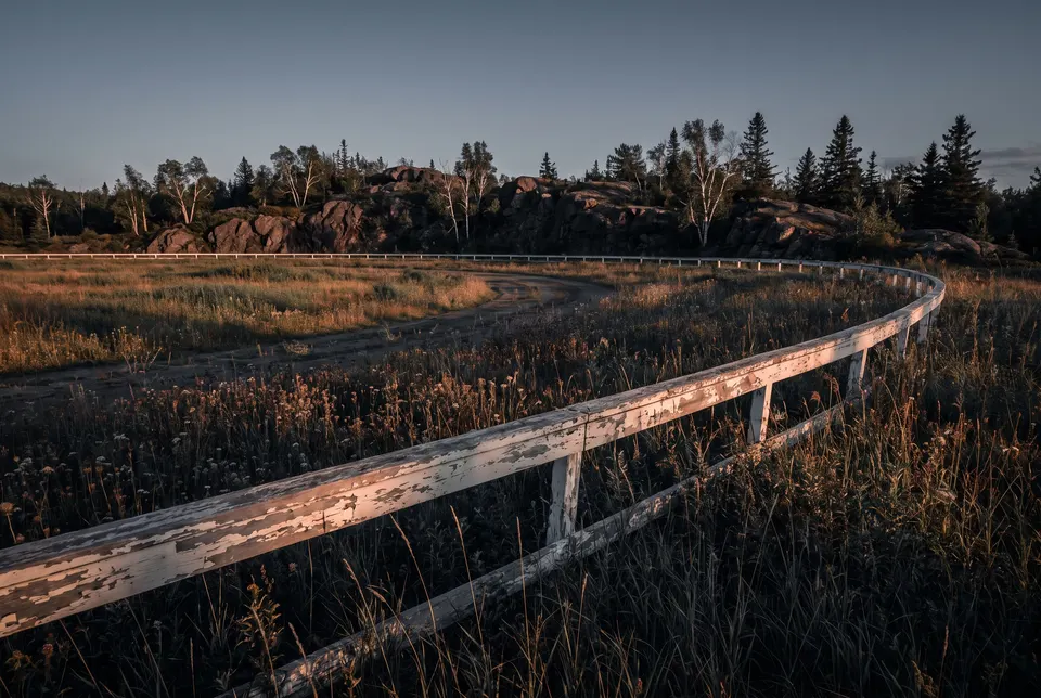 Generated scene: an old oval racetrack overgrown with grass, a weathered white rail curving in front of dark rock and birches at sunset.